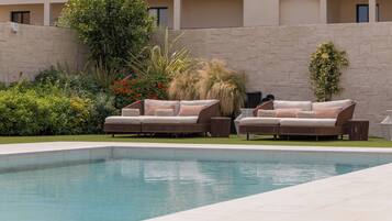 Zona de piscina con dos sofás beige y una mesa pequeña, rodeada de abundante vegetación y un muro de piedra.
