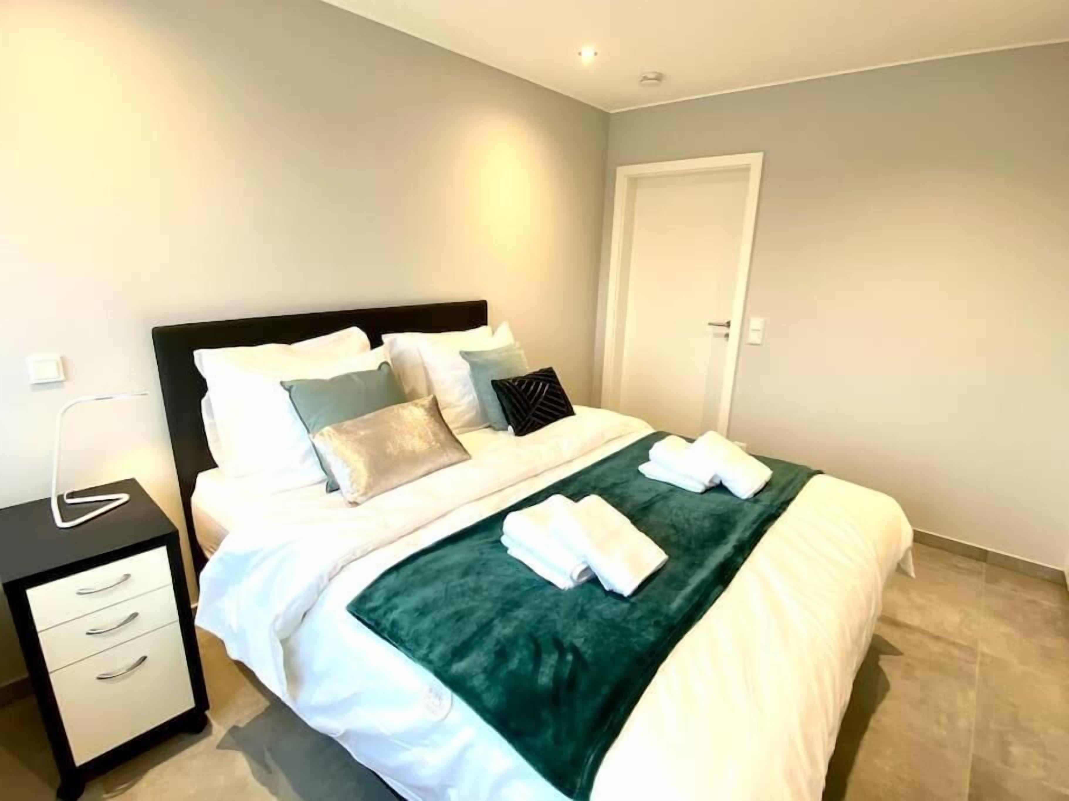 A neatly made bed with a green throw, white bedding, and several pillows. There is a bedside table with drawers and a lamp.