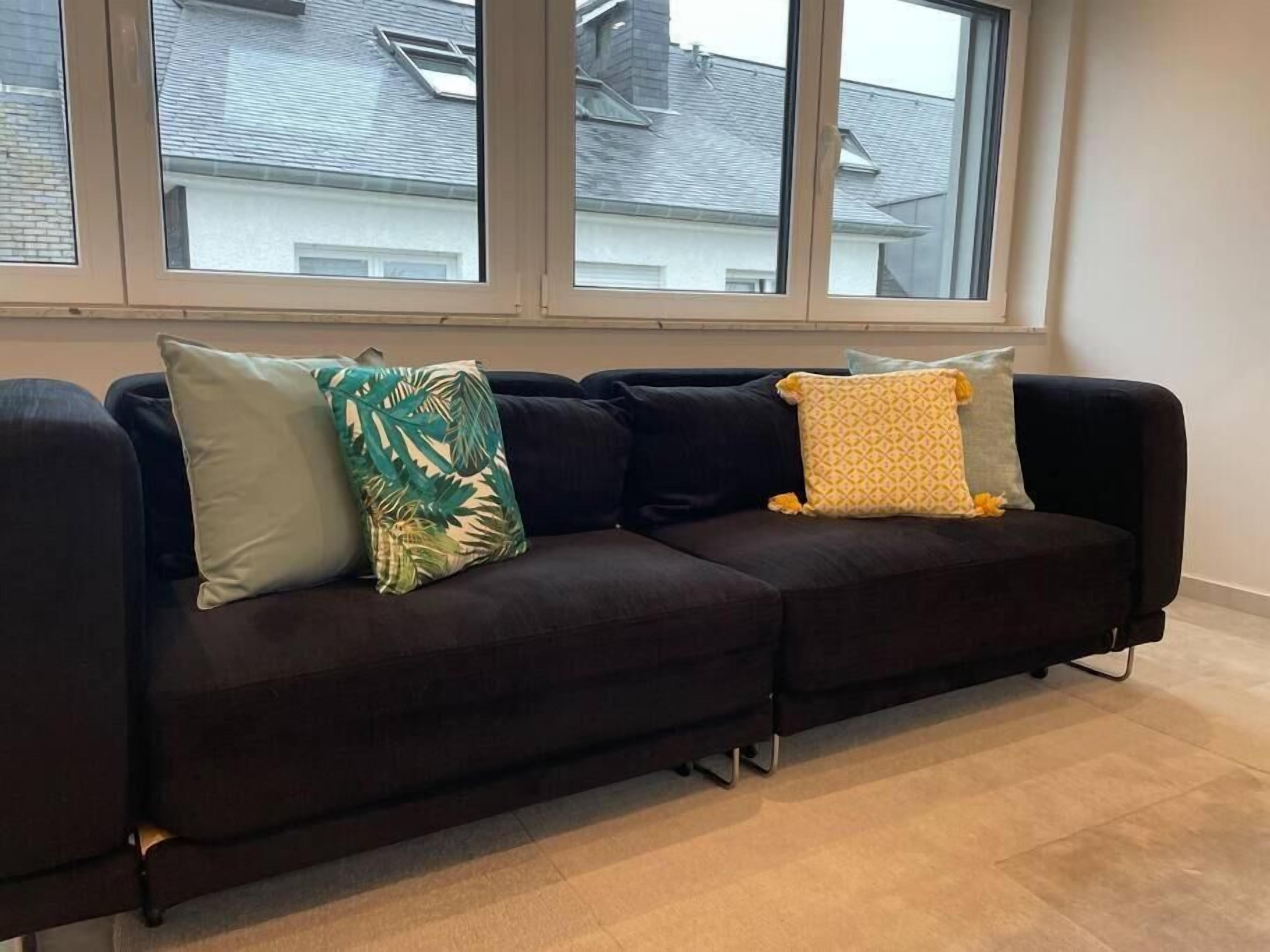 A dark-colored sofa with three cushions in front of a window with a view of neighboring houses.