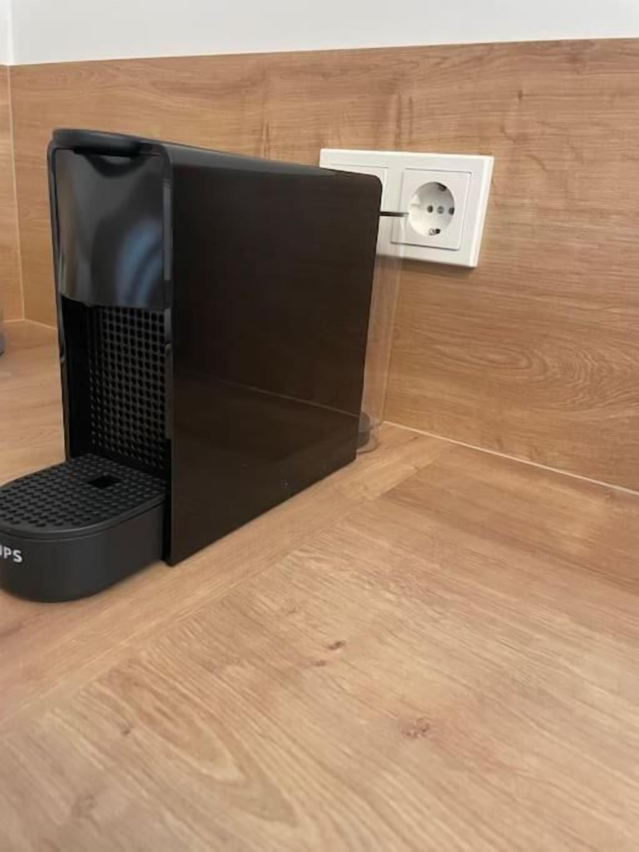 A black coffee machine with a drip tray on a wooden floor next to a wall socket.