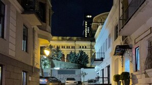 A narrow street at night with buildings on either side, a parked car, and a building with a clock tower in the background. - Opera Apartment Sleeps 4 With Patio (Tbilisi)