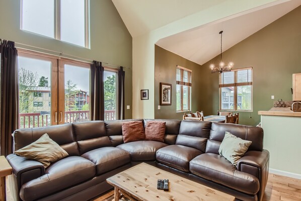 A spacious living room with a large L-shaped leather sofa, a wooden coffee table, and a view of the outside balcony.