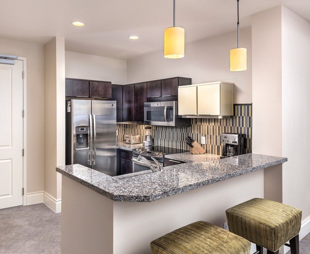 A modern kitchen with dark cabinets, stainless steel appliances, a granite countertop, and two woven stools.