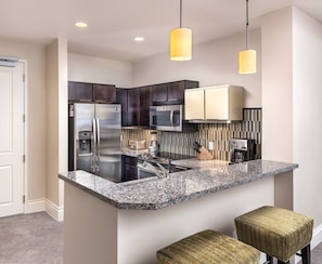A modern kitchen with dark cabinets, stainless steel appliances, a granite countertop, and two woven stools. - Las Vegas | Couples Skyline Lights & Strip Fun | Pool + Cabanas (Las Vegas)