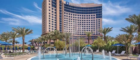 A hotel with a pool area featuring fountains, lounge chairs, and palm trees.
