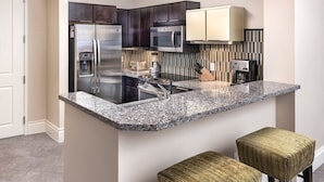 A modern kitchen with dark cabinets, stainless steel appliances, a granite countertop, and two woven stools. - Wyndham Desert Blue - 1 Bedroom Suite (Las Vegas)