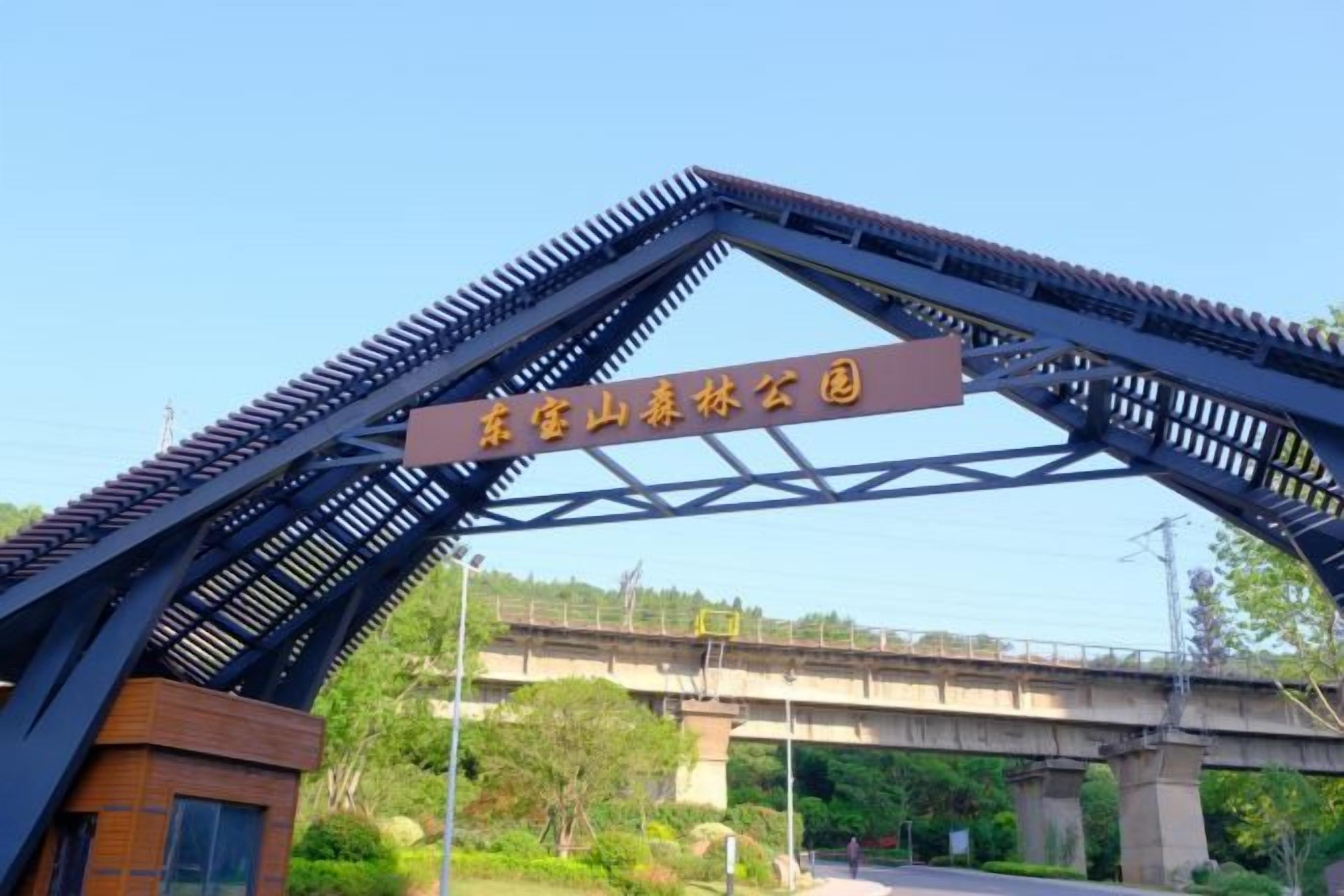 A metal archway with a sign, a bridge in the background, and greenery.