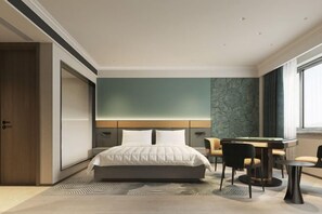 A modern hotel room with a large bed, a desk with chairs, and a view of the city.