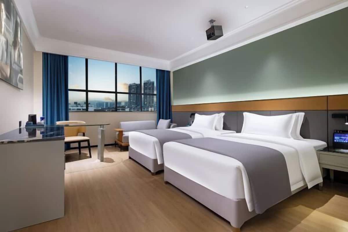 A modern hotel room with two beds, a desk, and a large window offering a city view.