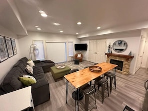A modern living room with a sectional sofa, a dining table with bar stools, a fireplace, and a flat-screen TV mounted on the wall.