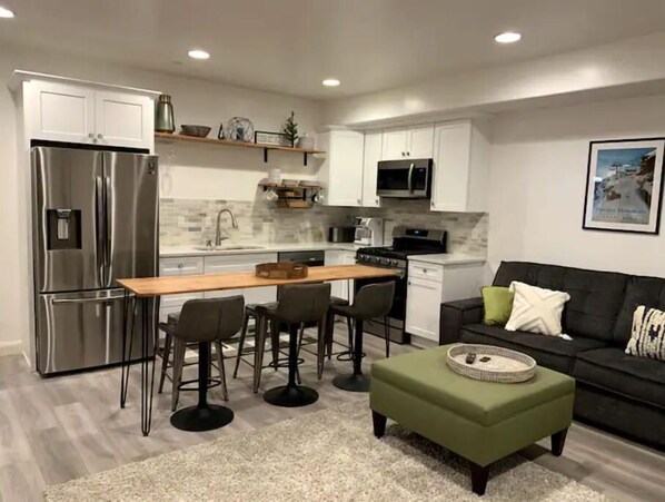 A modern kitchen with white cabinets, a stainless steel refrigerator, a microwave, and a stove. There is a dining area with a wooden table and four chairs. A green ottoman is placed in front of a black sofa with a throw pillow.