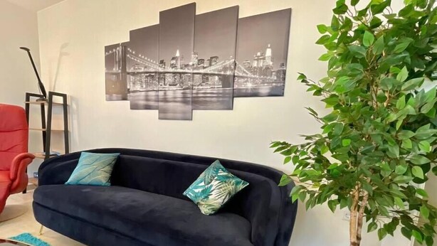Ein Wohnzimmer mit einem dunkelblauen Sofa, einem türkisgrünen Teppich, einer Topfpflanze und einem Stadtbild an der Wand.