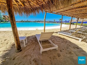 A beach with a thatched roof structure, white lounge chairs, and a pool in the background. - Bluu Habitat (Mazatlán)