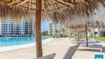 Zona junto a la piscina con sillones de descanso, techo de paja y un edificio al fondo.