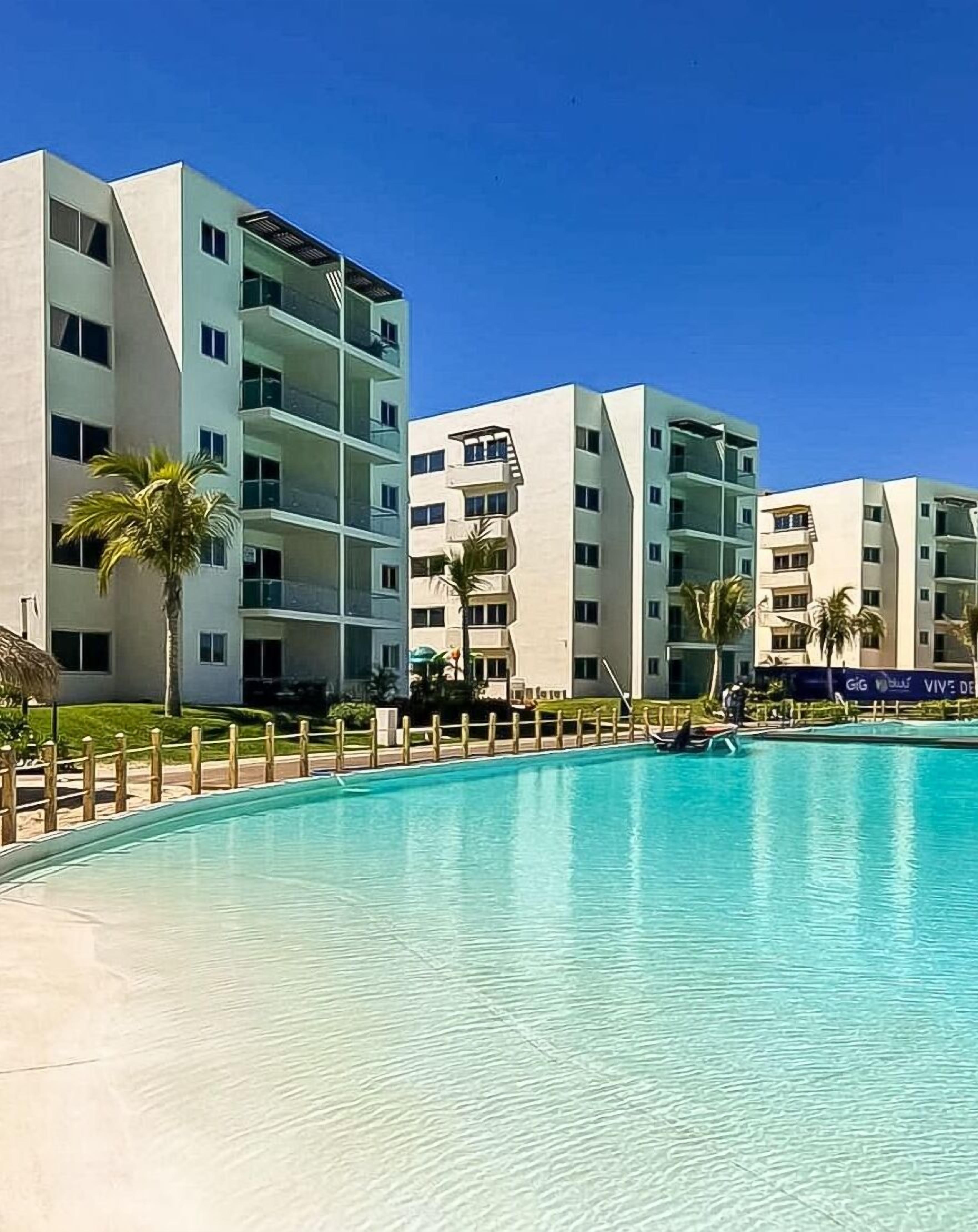 Een modern hotel met een zwembad, meerdere balkons en palmbomen.