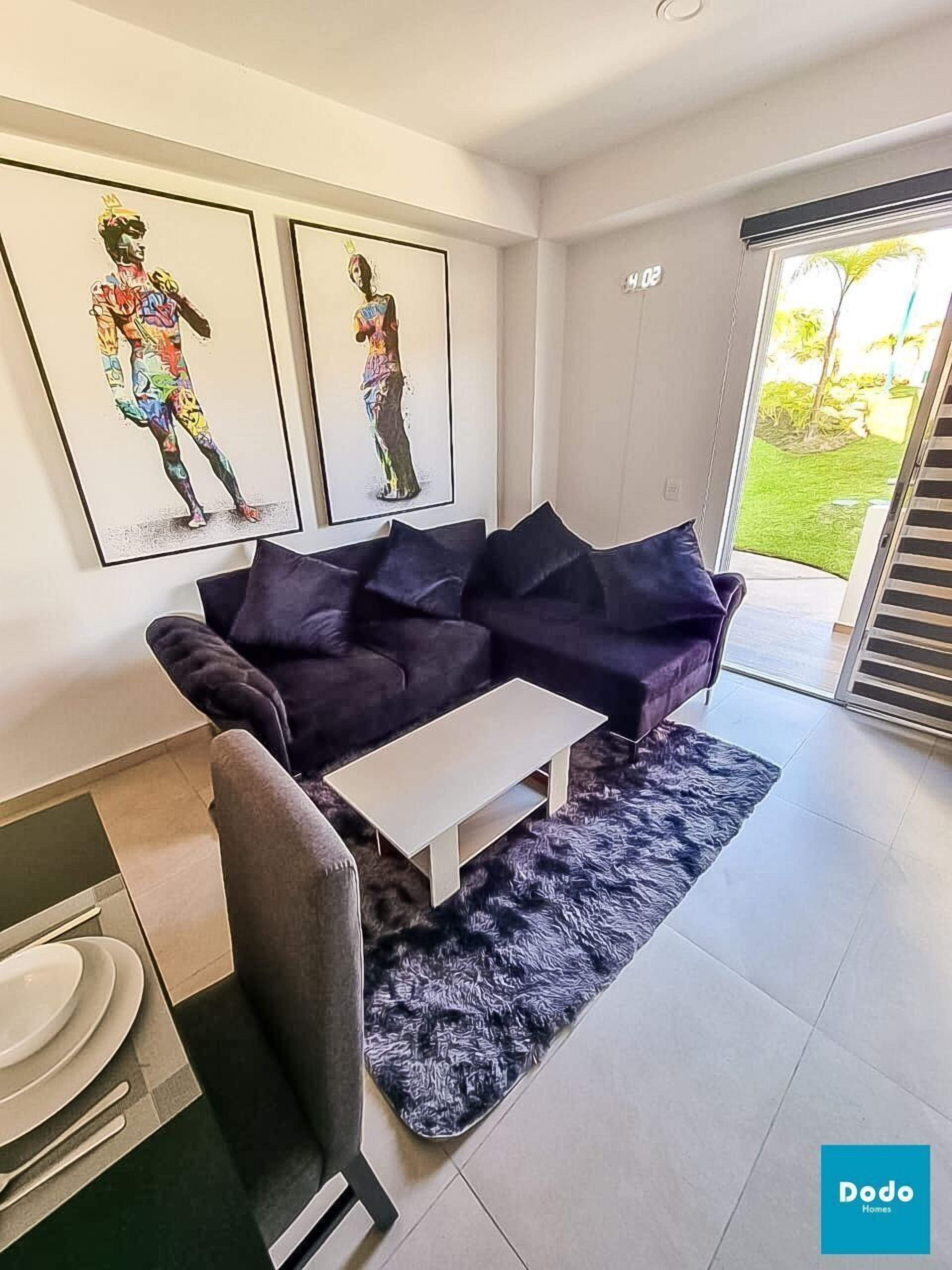 A modern living room with a dark purple sofa, a white coffee table, and a black furry rug. There are two framed artworks on the wall and a sliding glass door leading to a balcony.