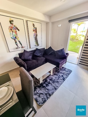 Un salon moderne avec un canapé de couleur violet foncé, une table basse blanche et un tapis noir à poils longs. Deux œuvres d’art encadrées ornent le mur, et une porte-fenêtre coulissante donne accès à un balcon.