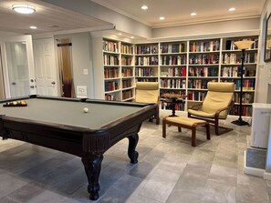 Une table de billard dans une pièce où se trouvent une bibliothèque et deux chaises.