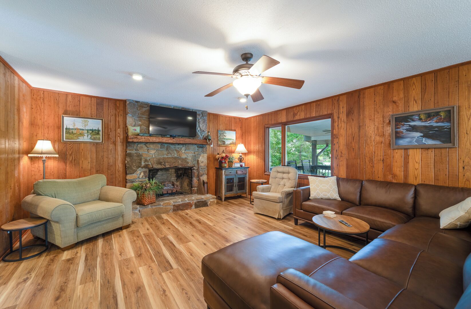 A cozy living room with wooden walls, a stone fireplace, a ceiling fan, and a large window.