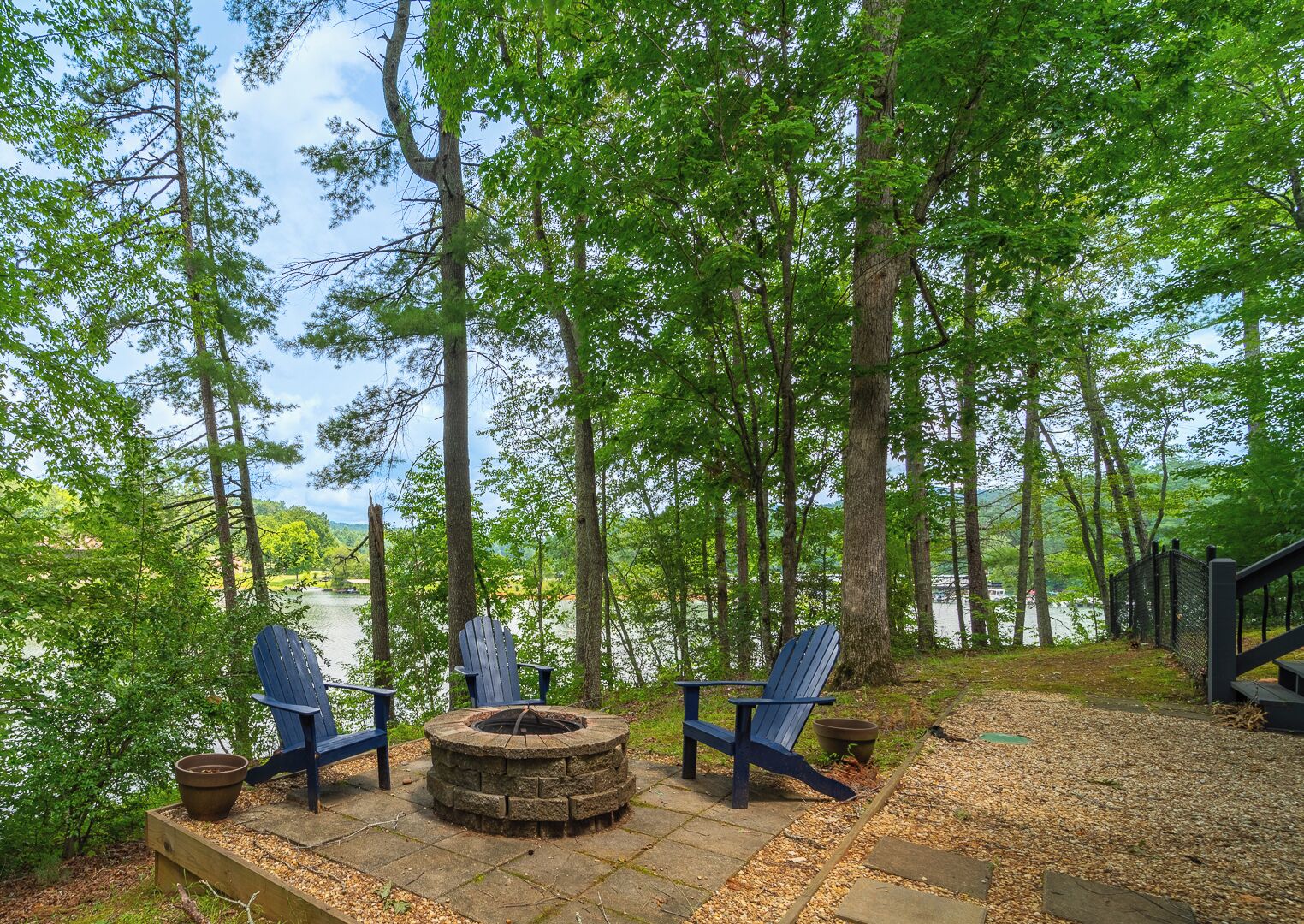 Een bebost gebied met een vuurplaats van natuursteen en twee Adirondack-stoelen gericht op een meer.
