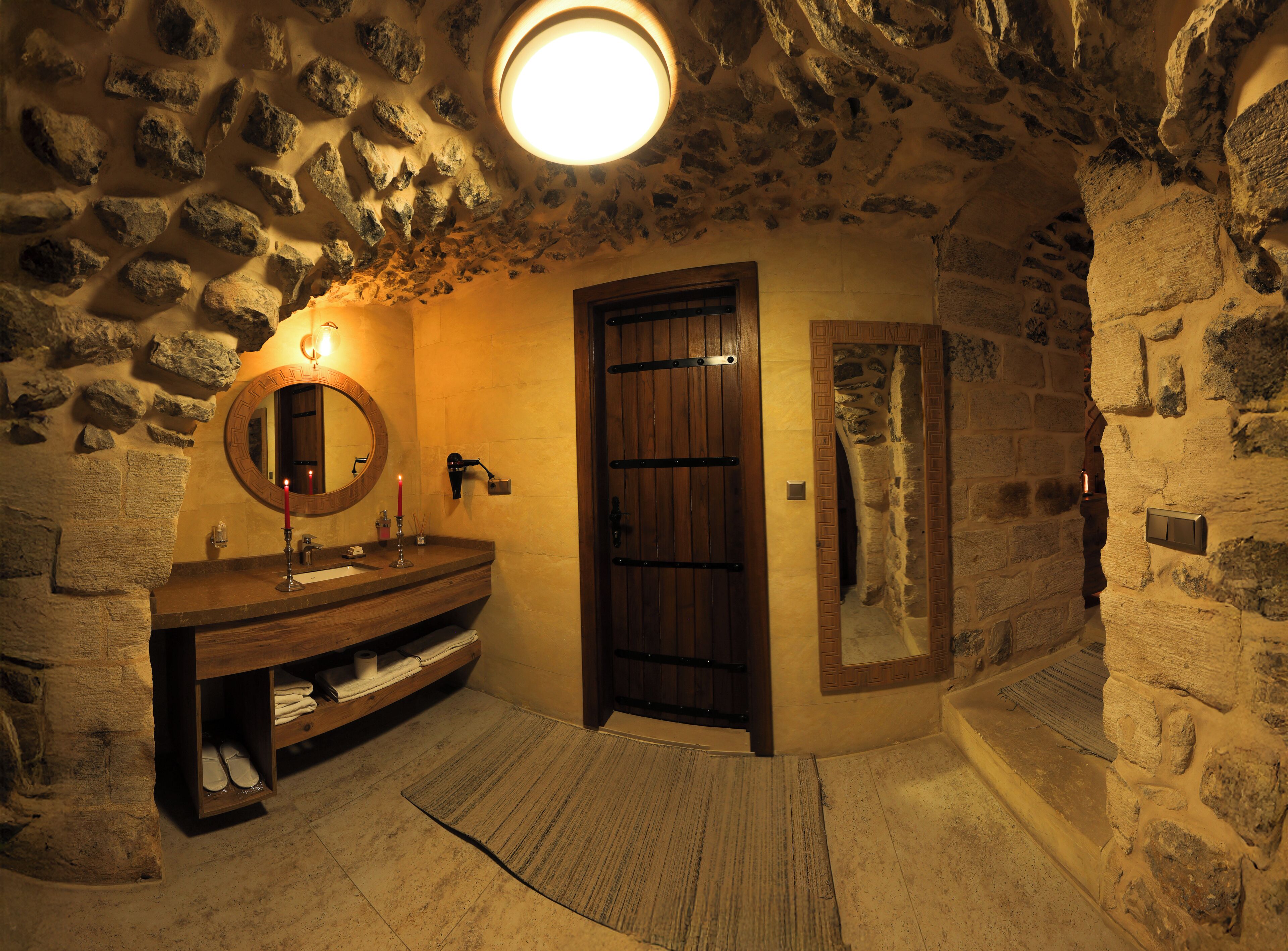A stone-walled bathroom with a wooden vanity, a round mirror, a lit candle, and a door leading to another room.