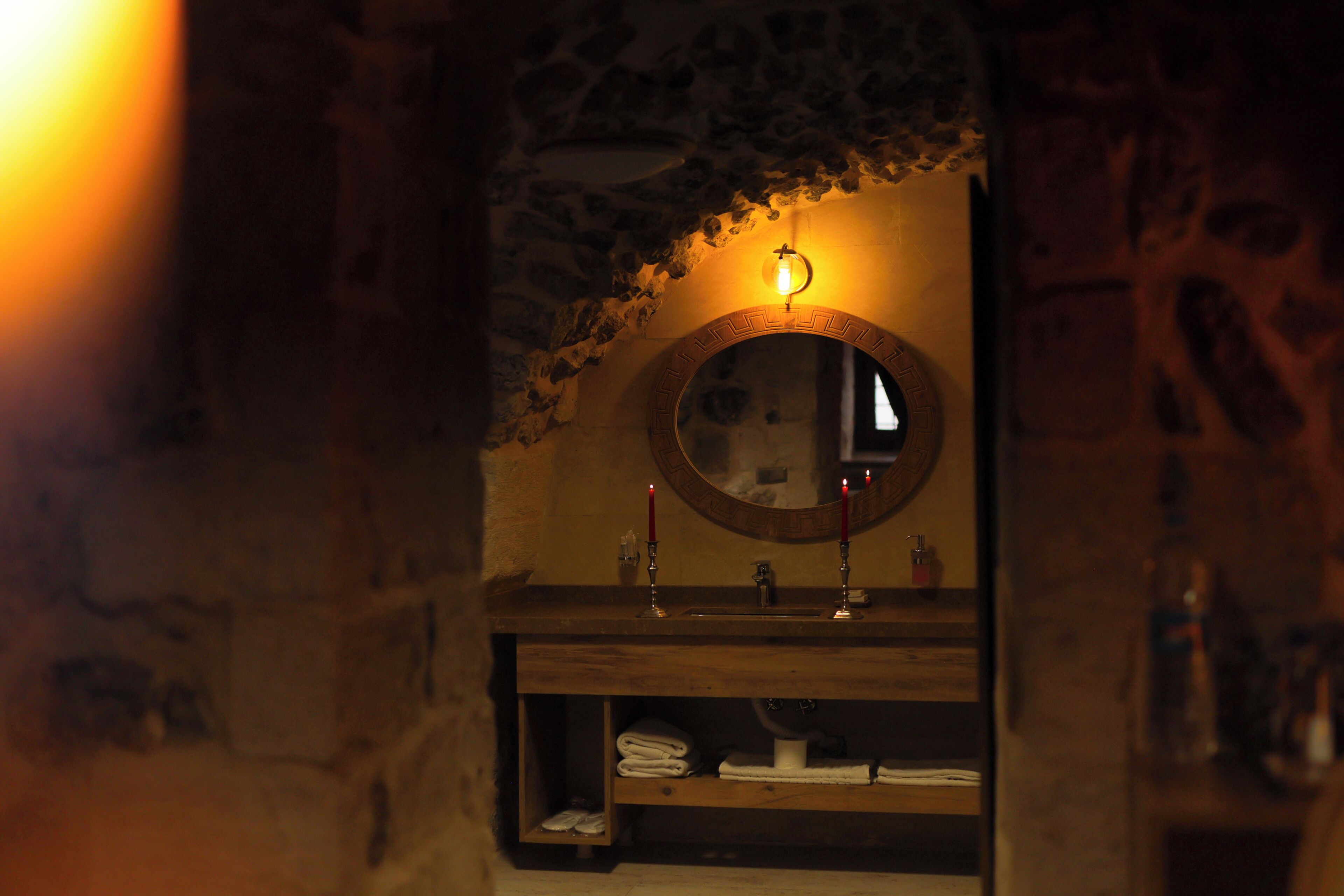 A stone-walled bathroom with a wooden vanity, a round mirror, and a shelf with folded towels.