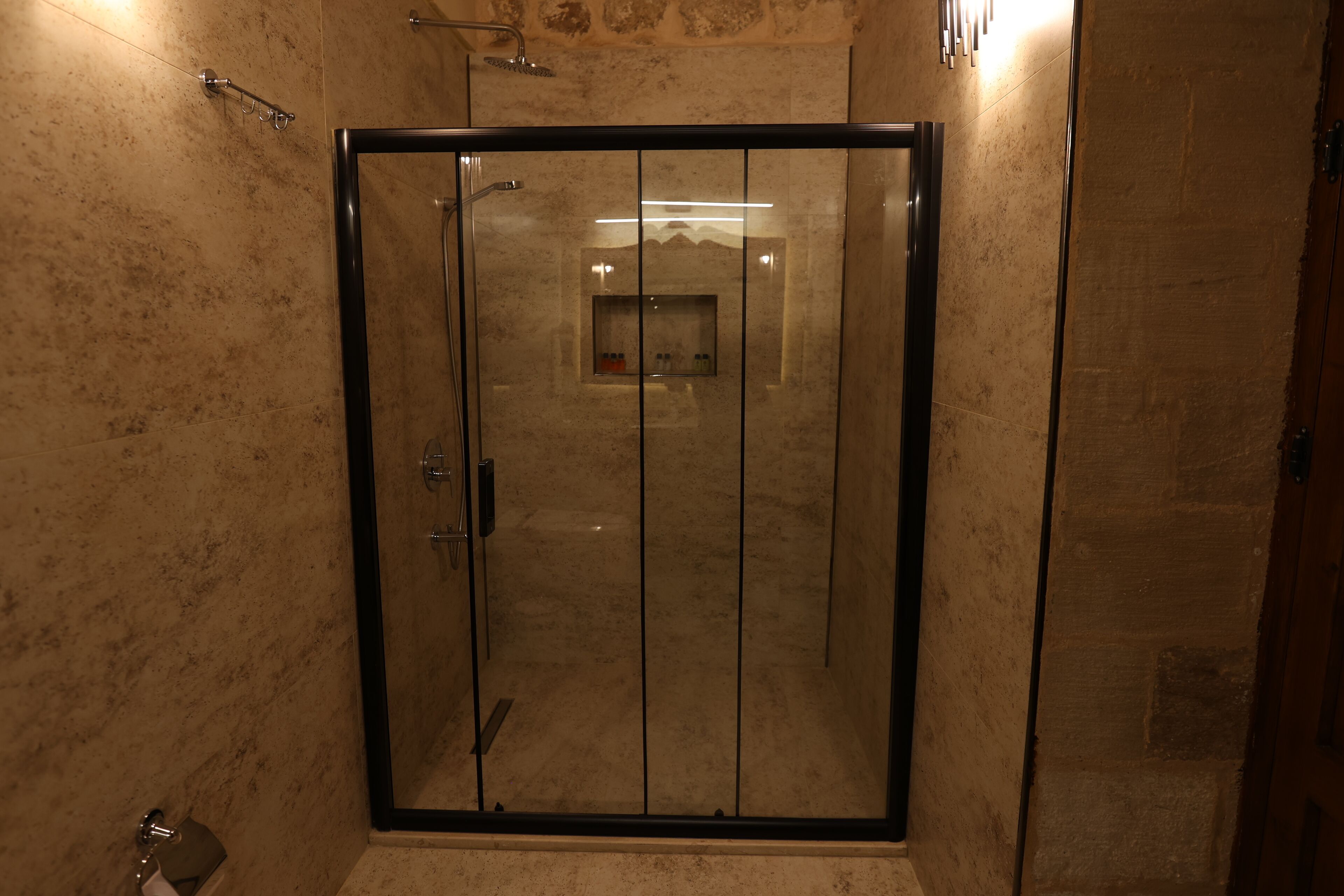 A glass-enclosed shower with a marble wall and a small shelf with toiletries.