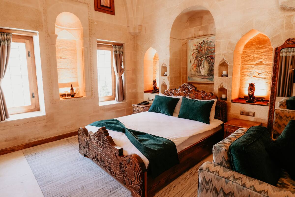 Ein Schlafzimmer mit einem großen Bett, einem Holz-Kopfstück und einem Nachttisch mit Lampe. Zwei Fenster mit Vorhängen, ein Bild an der Wand und ein Spiegel mit dekorativem Rahmen sind ebenfalls vorhanden.