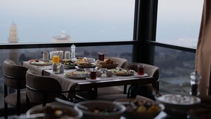 Una mesa de comedor dispuesta con diferentes platos y bebidas, con vistas a un paisaje urbano donde destaca una torre imponente.
