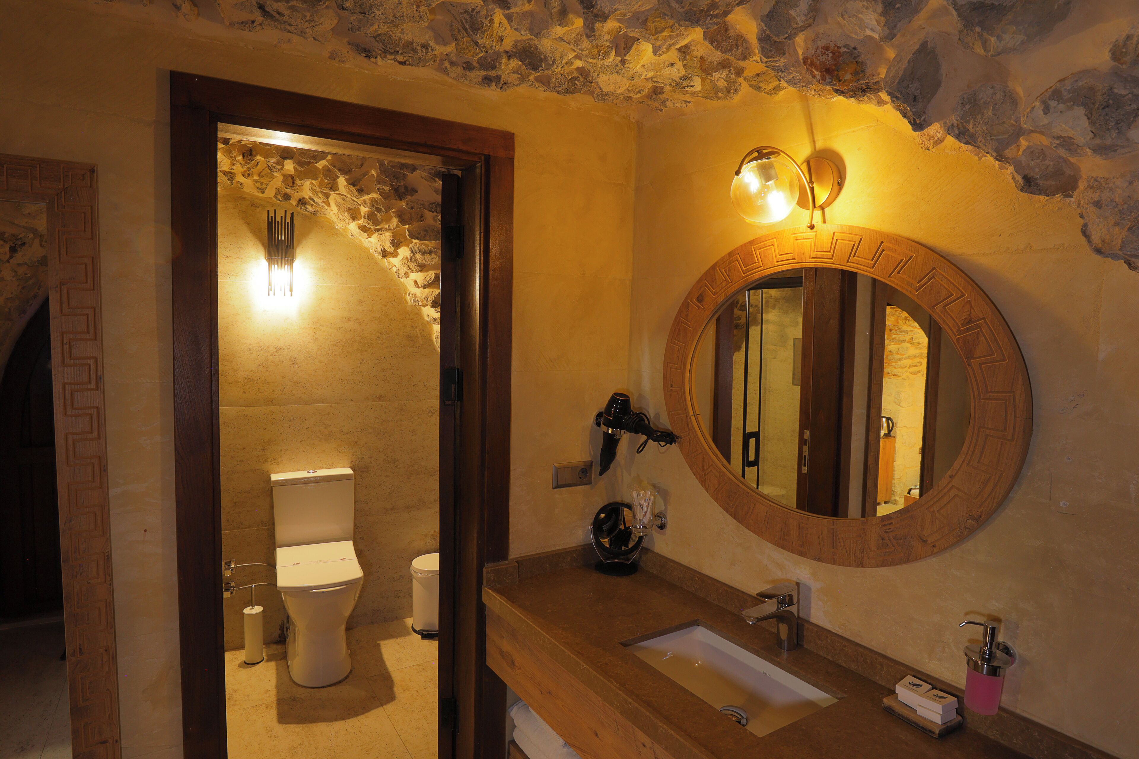 A bathroom with a stone wall, a wooden vanity, a round mirror, and a toilet.