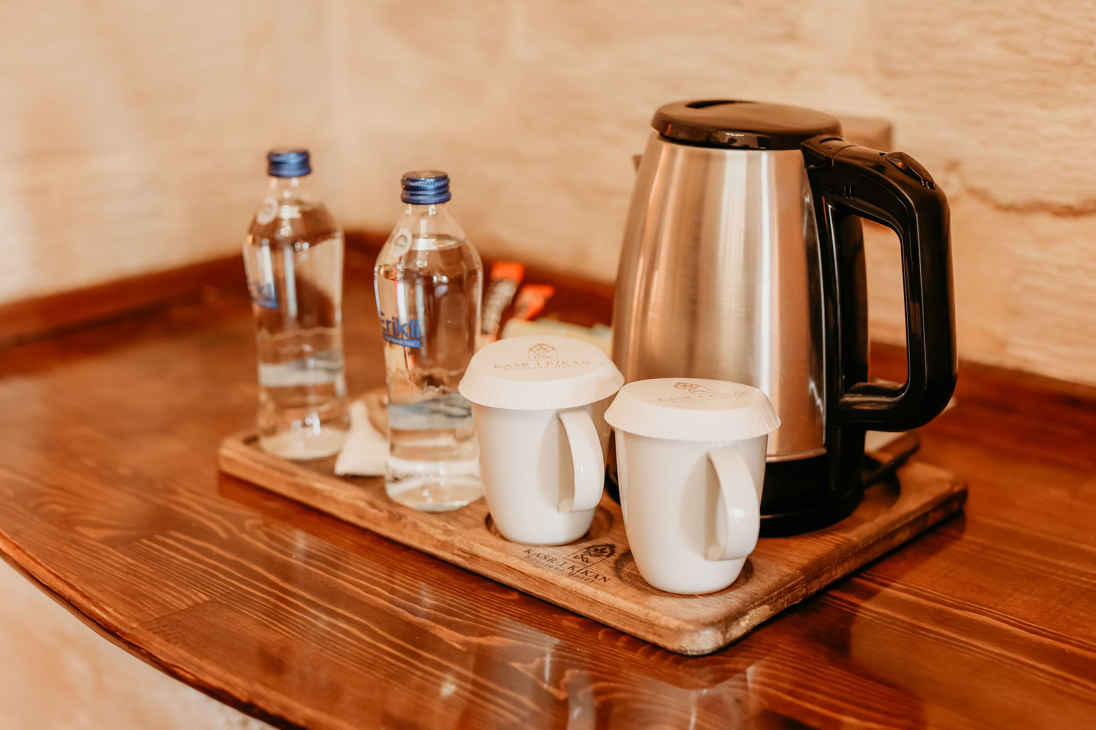 A wooden tray with a stainless steel coffee pot, two white mugs, and two water bottles on a wooden table.