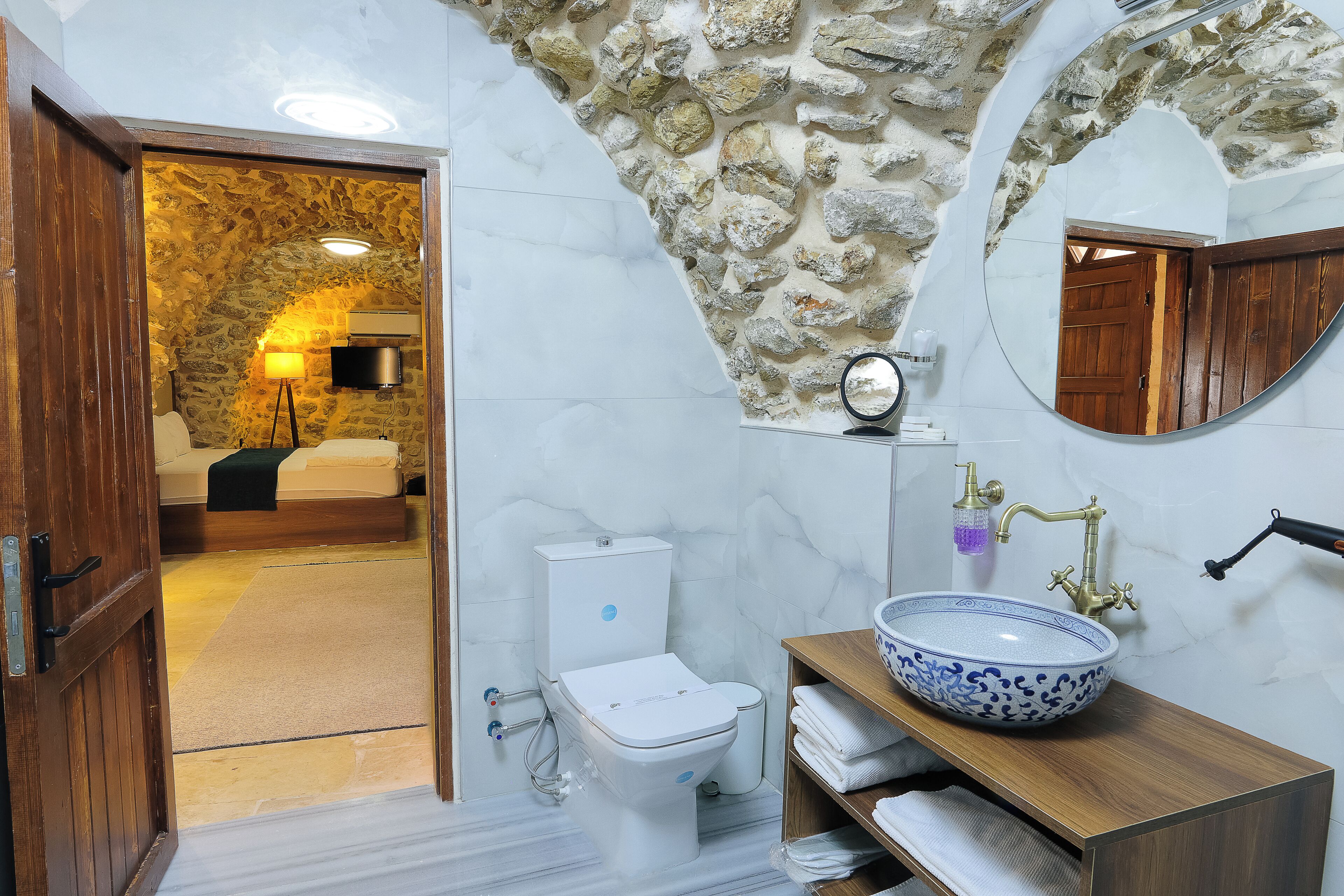 A bathroom with a stone wall, a white toilet, a blue and white bowl sink, a wooden vanity, and a mirror.