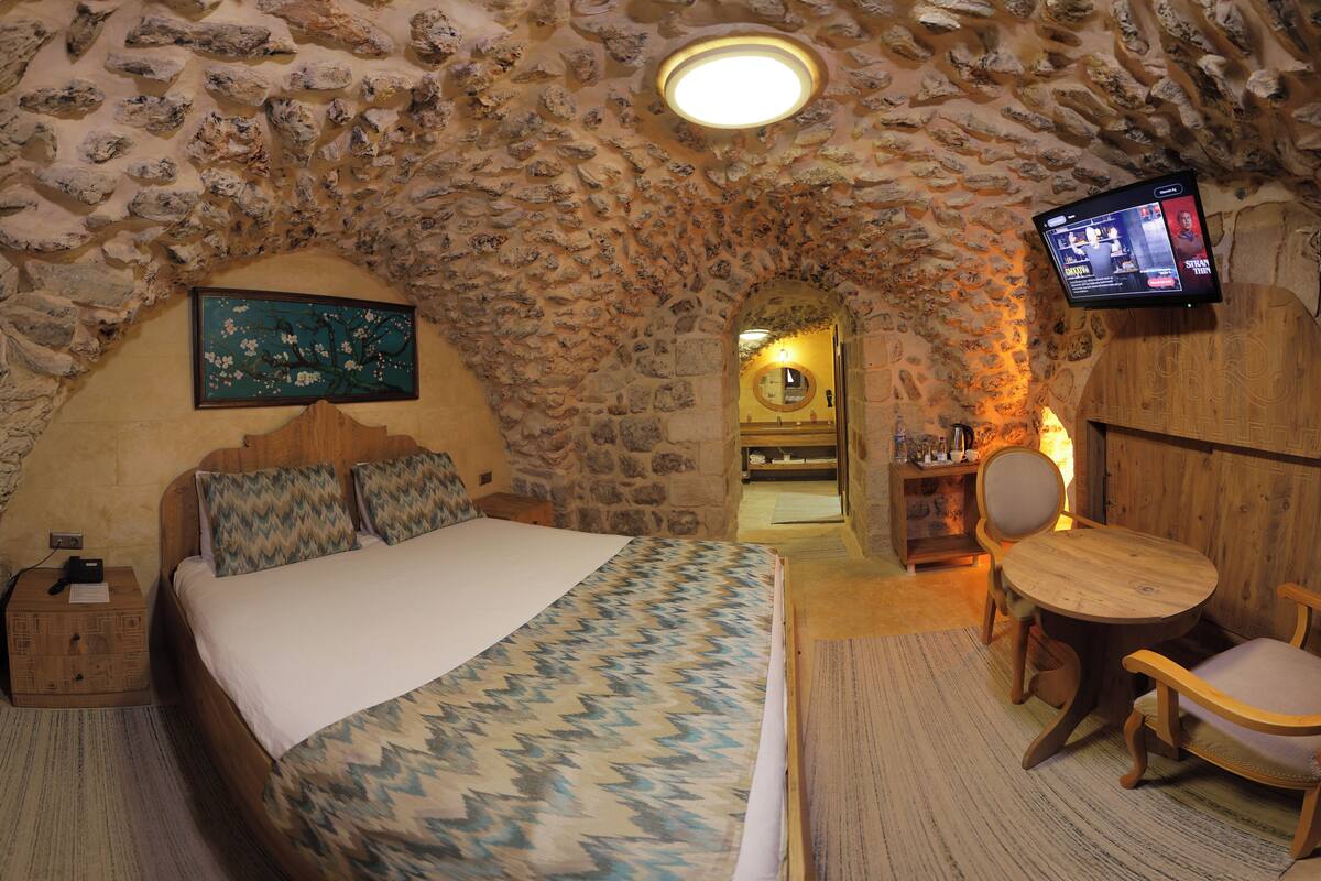 Ein gemütliches Zimmer mit Steindecke, einem Bett mit gemusterten Betttextilien, einem Holztisch und einem an der Wand befestigten Fernseher.