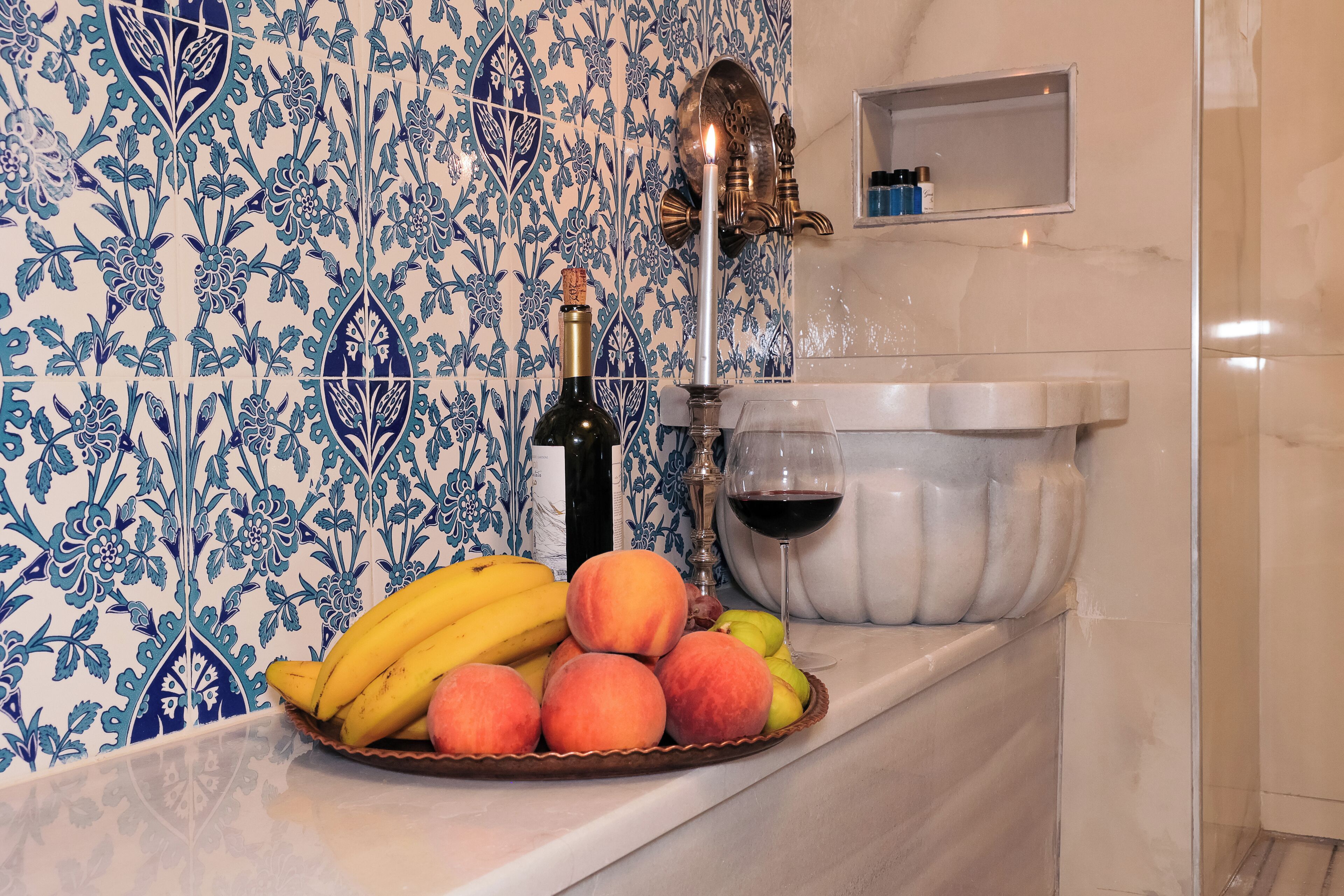 A kitchen counter with a fruit bowl containing bananas, peaches, and apples, a bottle of wine, and a wine glass.