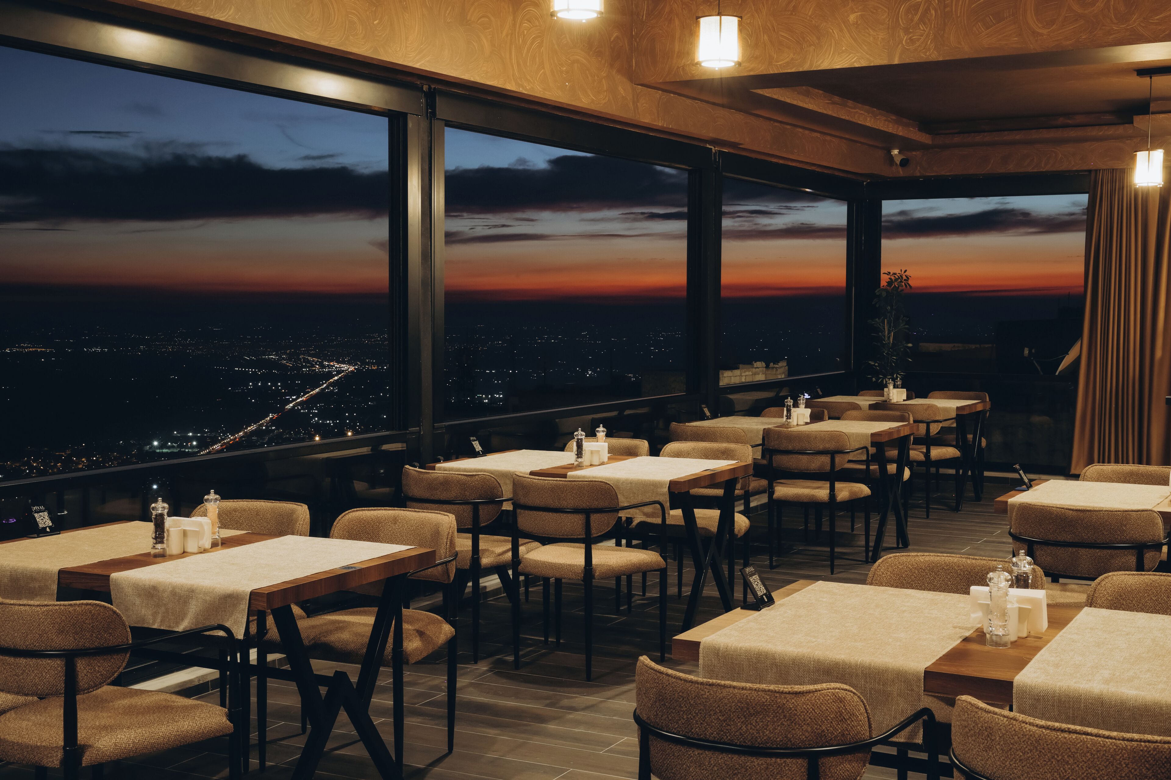 Un restaurante con mesas y sillas dispuestas para comer, amplios ventanales que ofrecen vistas del paisaje urbano al atardecer.