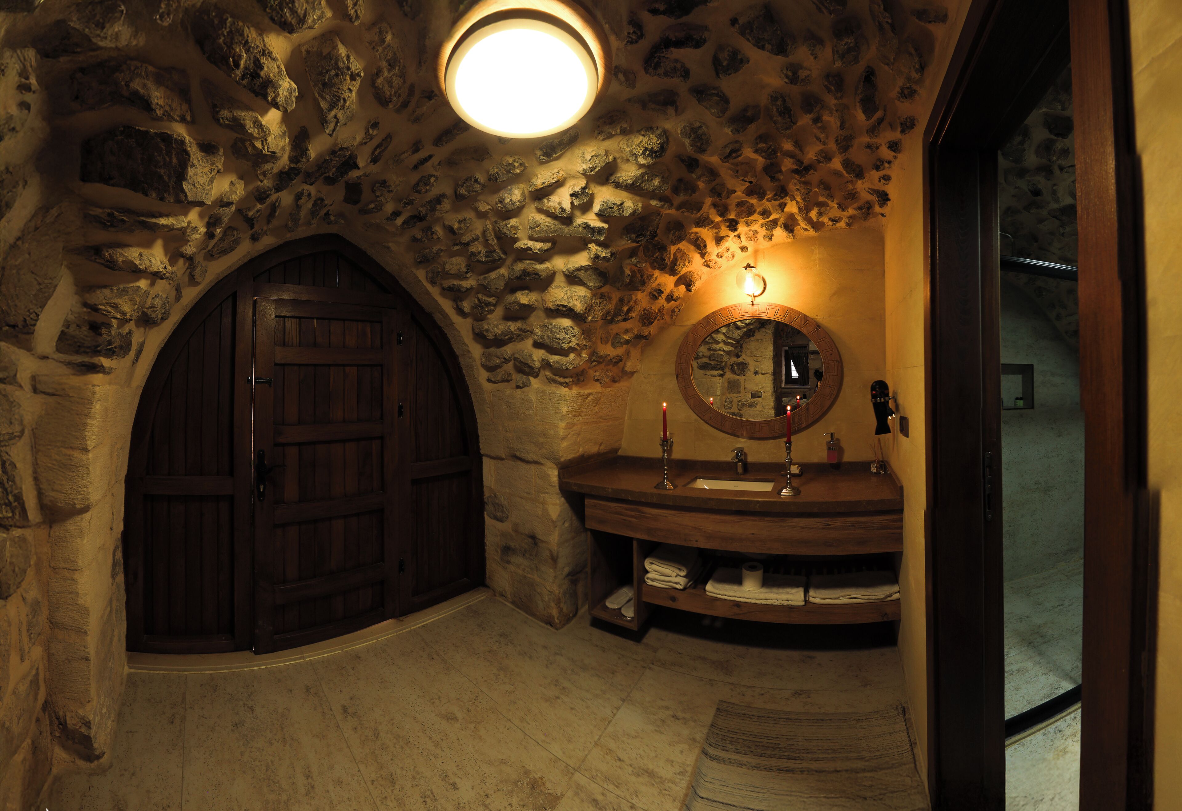 A stone-walled bathroom with a wooden door, a round mirror, and a sink with a towel rack.