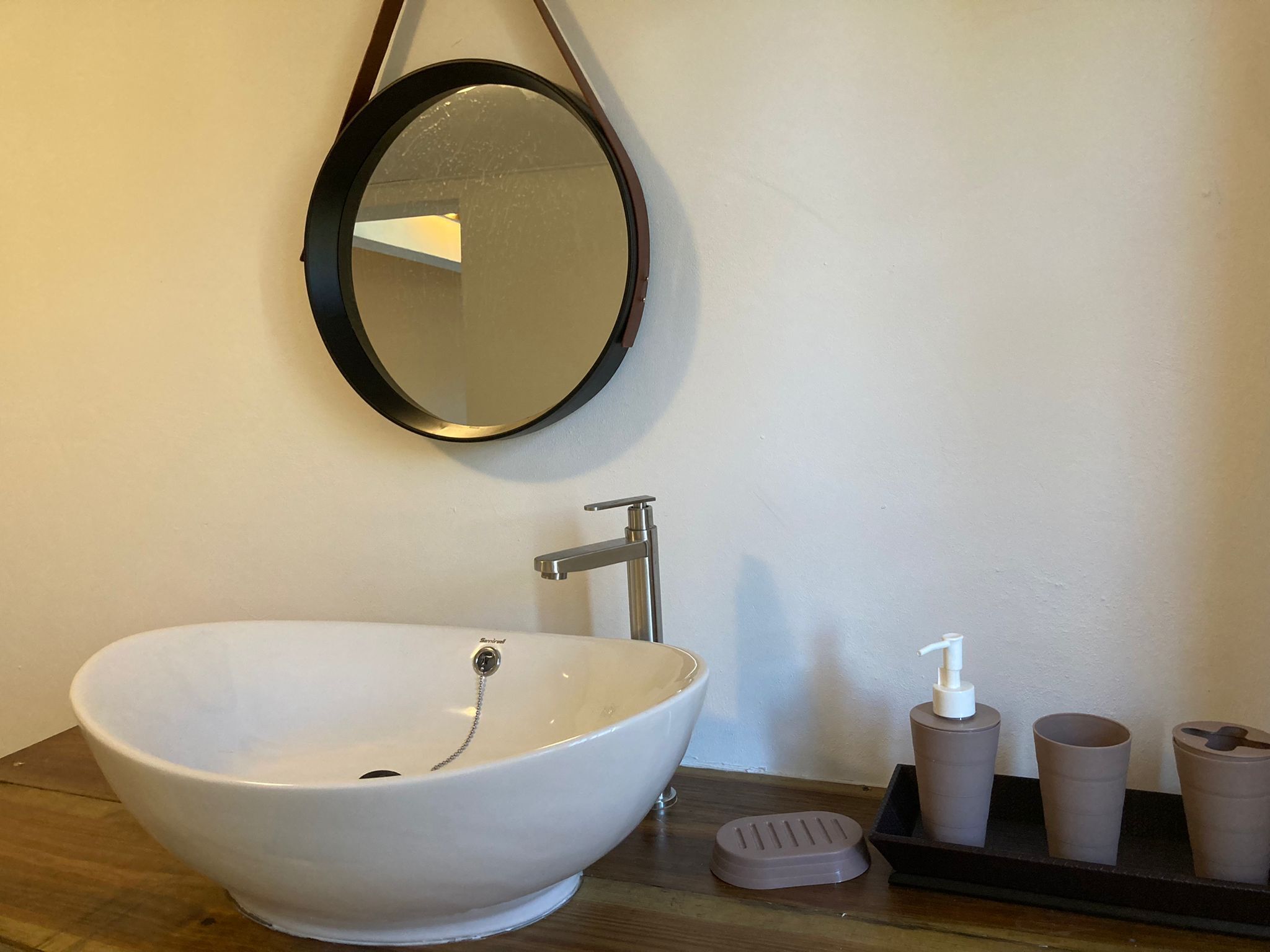 Un baño con un lavamanos blanco de diseño, un grifo moderno, un espejo redondo y una pequeña bandeja con artículos de aseo.