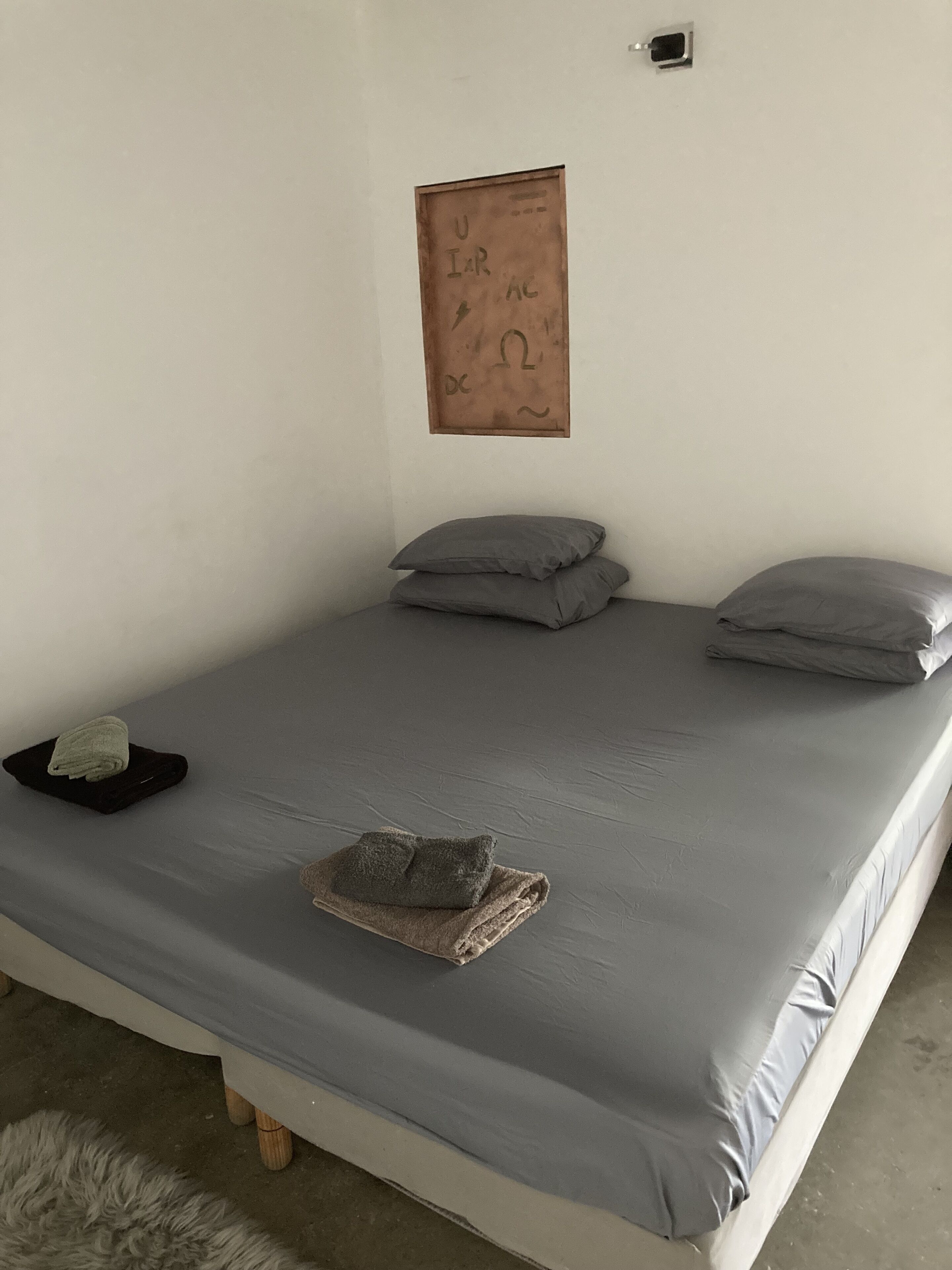A single bed with grey bedding, two pillows, a folded towel, and a small framed item on the wall.