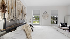 Un dormitorio con una cama grande, un tapiz de macramé y vistas al exterior a través de dos ventanas.