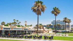 Zona esterna con tavoli e sedie, palme e un edificio con insegna 'mana'.