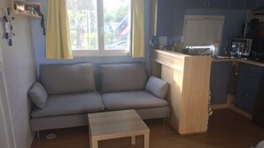 A compact living space with a grey sofa, a wooden coffee table, and a kitchenette area. - Mobil Home/bungalow 6 Places Camping 4 (Hyères)