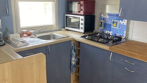 A compact kitchen with blue cabinets, a gas stove, a microwave, and a window. - Mobil Home/bungalow 6 Places Camping 4 (Hyères)