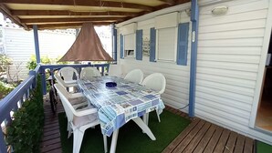A covered patio with a table and chairs, a small grill, and a white mobile home with blue shutters. - Mobil Home/bungalow 6 Places Camping 4 (Hyères)