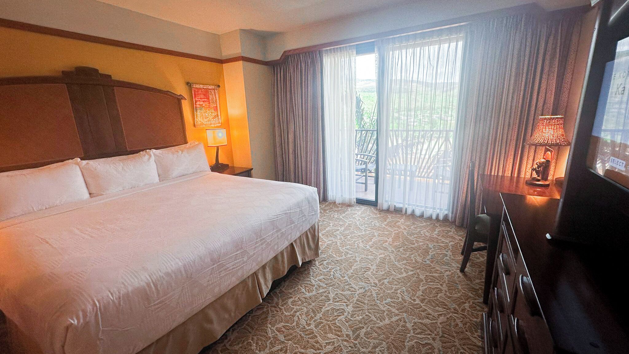 A hotel room with a large bed, a desk, a chair, a lamp, and a view of the outdoors through the window.