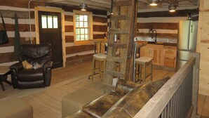 A rustic cabin interior with a wooden chair, a ladder, a bar area, and a kitchen with a refrigerator.