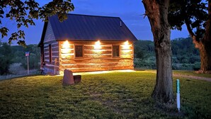 A log cabin with a metal roof, surrounded by a grassy area and a large tree.
