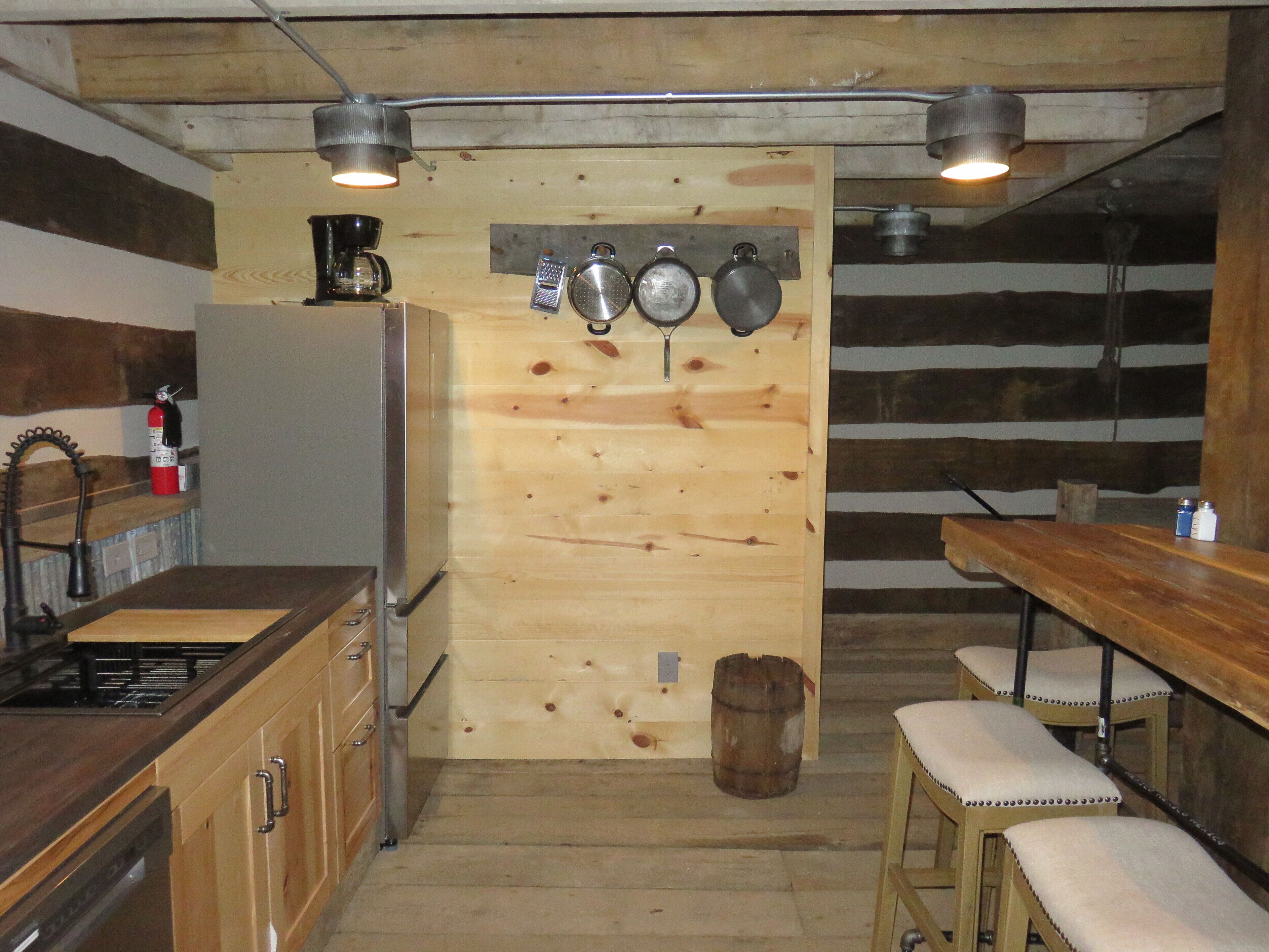 A rustic kitchen with wooden walls, a wooden table, and a bar stool. There are hanging pots and a fire extinguisher on the wall.