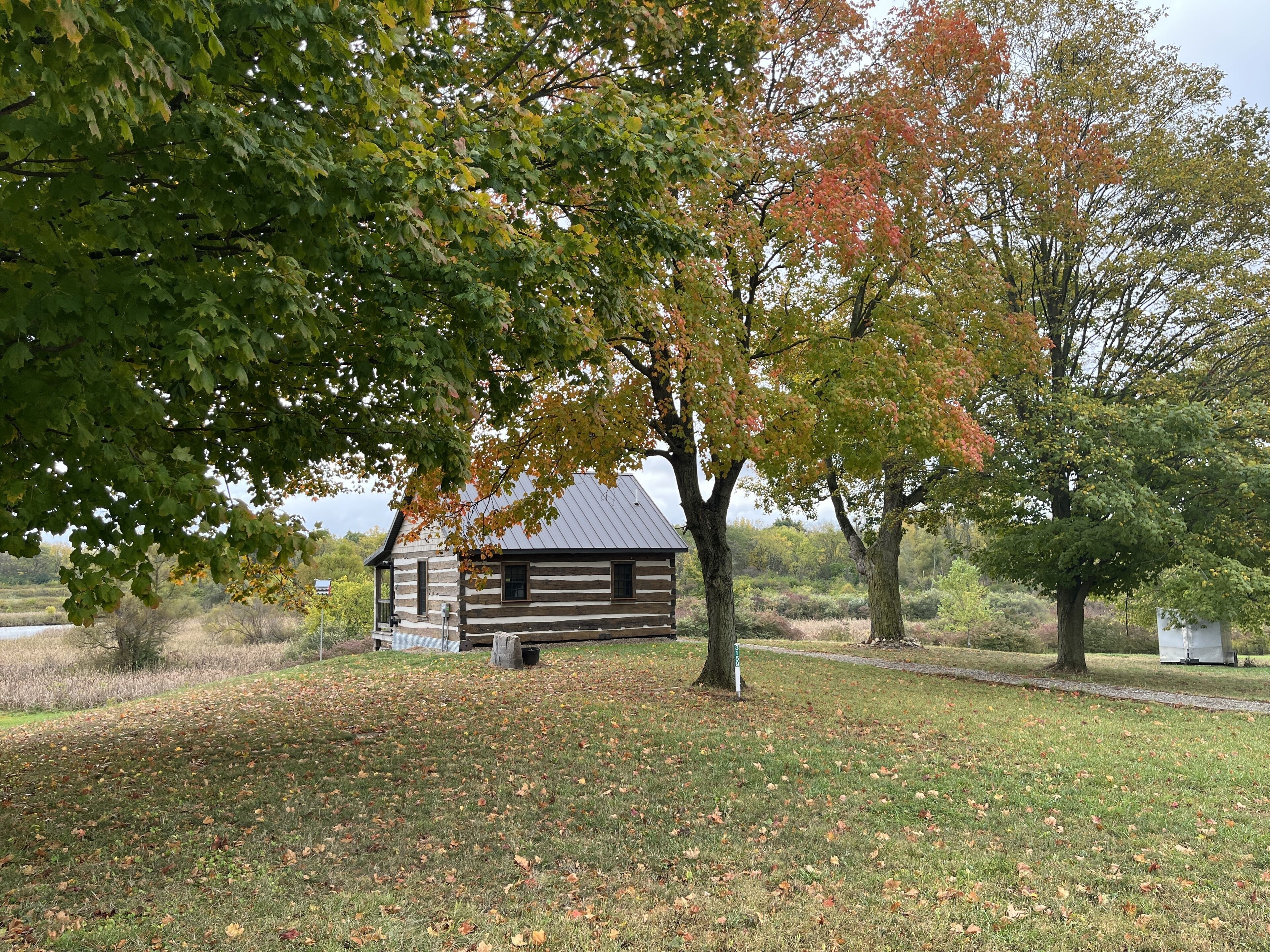 Un chalet rustique en rondins, entouré d’arbres aux feuilles d’automne, situé dans une zone herbeuse avec un étang en arrière-plan.