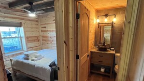 A wooden cabin bedroom with a bed, a small bathroom with a mirror and sink, and a window.
