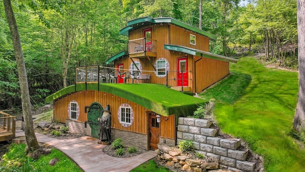 Una cabaña de madera de dos pisos con techo verde, rodeada de abundante vegetación y árboles.